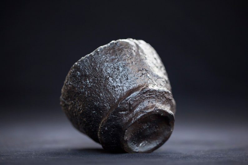 K&ouml;nnte beinhalten: Eine dunkelgraue Teetasse aus Keramik mit einer rauen, strukturierten Oberfl&auml;che. Die Tasse ist leicht geneigt und scheint handgefertigt zu sein.