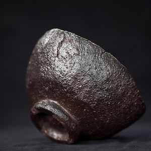 May include: A dark brown, textured ceramic bowl with a rough, uneven surface. The bowl has a small, rounded base and a slightly irregular rim. The overall shape is organic and asymmetrical, suggesting a handmade quality.