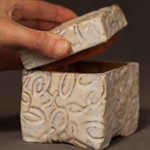 Peut inclure: Une boîte carrée en céramique blanc cassé avec un couvercle soulevé. La boîte et le couvercle sont texturés avec un motif floral répétitif. L'intérieur de la boîte est de couleur marron chaud.