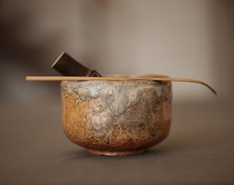 Puede incluir: Un cuenco de té de cerámica artesanal con una superficie texturizada y moteada en tonos marrones y grises. Un batidor de bambú y una cuchara de madera descansan en el borde, sugiriendo una ceremonia del té.