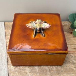May include: A square ceramic box with a glossy amber-colored glaze. The lid features a three-dimensional sculpted bee with yellow, brown, and white painted details. The box has a smooth, reflective finish and a flat, square-shaped lid.