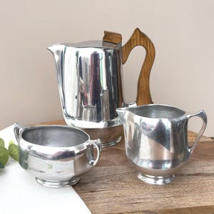 Puede incluir: Un juego de té de tres piezas de metal plateado que consta de una cafetera alta con mango de madera, una lechera y un azucarero. Cada pieza presenta un acabado pulido y reflectante con un diseño suave y redondeado con asas integradas.