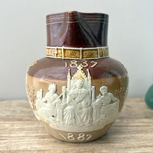 May include: A two-toned brown and tan ceramic jug featuring a relief design of a monarch seated on a throne flanked by two figures. The piece displays the years 1837 and 1887 in raised white numerals and includes a belt-like band detail.