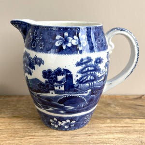 May include: A ceramic pitcher featuring a blue and white transferware pattern. The design displays a landscape with a bridge, buildings, and trees, bordered by floral motifs at the top and bottom. It has a curved handle and a pouring spout.