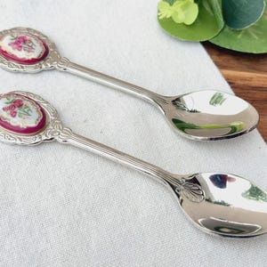 May include: Two silver-toned metal spoons featuring an oval-shaped decorative inlay on the handle. The inlay is white with a pink floral design, bordered by a deep magenta rim and an ornate, embossed silver-toned metal frame.