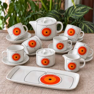 May include: A white ceramic tea set featuring a repeating graphic pattern of black-outlined squares and concentric orange and red circles with a black centre. The set includes a teapot, a sugar bowl, a creamer, six cups, six saucers, and a rectangular tray.
