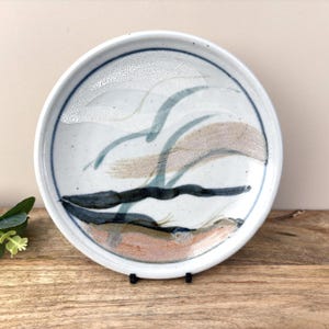 Puede incluir: Un plato de cerámica blanco con un diseño abstracto pintado a mano. El diseño presenta pinceladas azules, beige y negras, creando una composición similar a un paisaje. El plato tiene un borde azul oscuro y se exhibe sobre una superficie de madera.