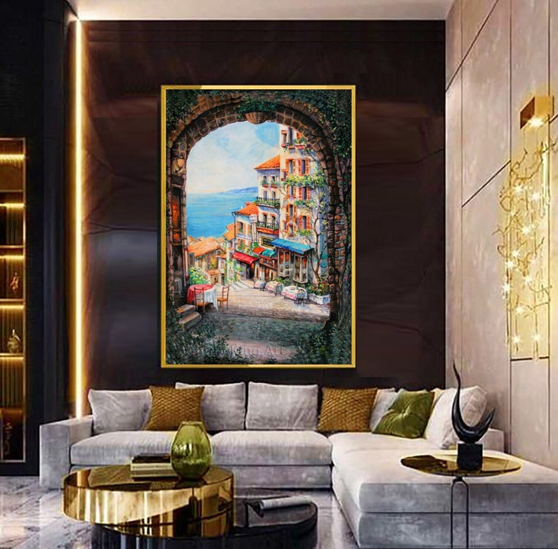 May include: A painting of a colorful cityscape with buildings, a stone archway, and a view of the ocean. The painting is framed in gold and hangs on a dark wall.