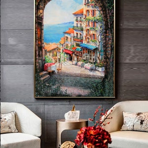 May include: A painting of a colorful, European-style town with a view of the ocean. The painting is framed in a gold frame and is hanging on a wall. The town is seen through an archway, with buildings, shops, and people walking around.