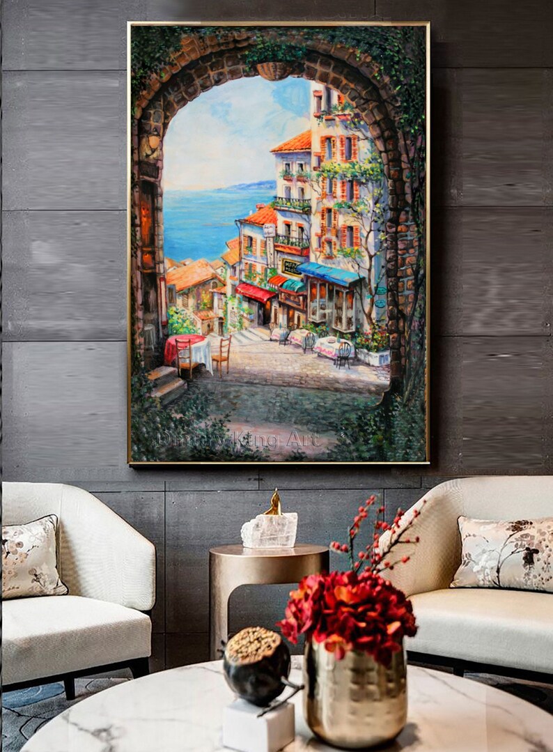 May include: A painting of a colorful, European-style town with a view of the ocean. The painting is framed in a gold frame and is hanging on a wall. The town is seen through an archway, with buildings, shops, and people walking around.