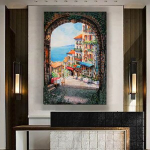 May include: A painting of a colorful, European cityscape viewed through a stone archway. The scene features buildings with red tile roofs, balconies, and lush greenery. The ocean is visible in the distance.