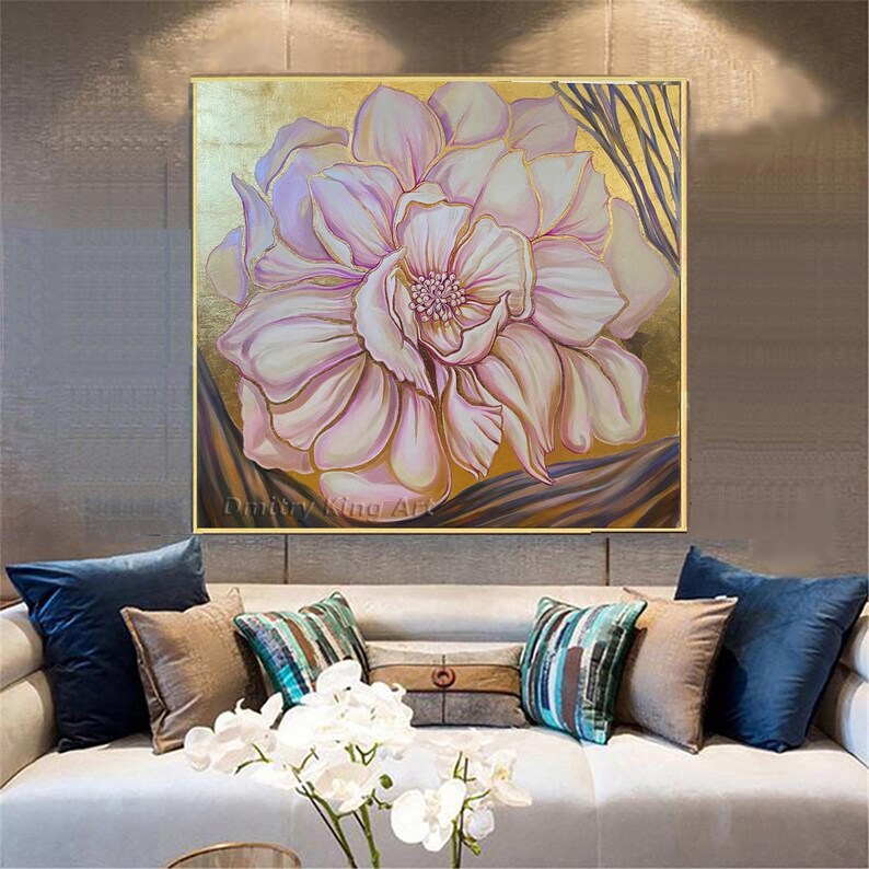 May include: A large, pink flower with a white center is painted on a canvas with gold leaf accents. The painting is framed in a gold frame and hangs on a wall.