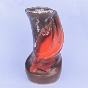 May include: A small, abstract ceramic vase with a brown, orange and white glaze. The vase has a unique, asymmetrical shape.