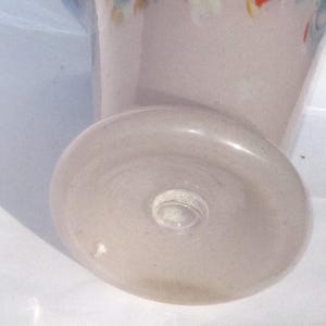 May include: A light pink glass vase with a rounded base and a flared top. The base is approximately 3 inches in diameter. The vase has a clear glass stem and a decorative rim with colorful accents.