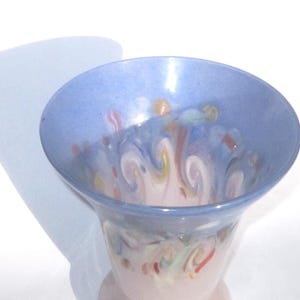 May include: A decorative glass vase with a flared rim and a light pink base. The vase features a light blue rim and interior with colorful, swirled patterns in shades of pink, yellow, and blue. The vase is set against a white background.