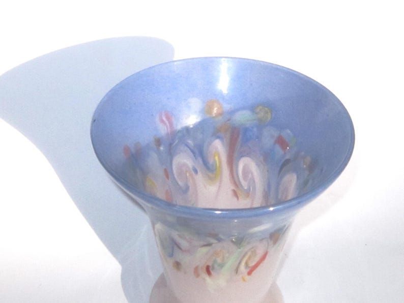 May include: A decorative glass vase with a flared rim and a light pink base. The vase features a light blue rim and interior with colorful, swirled patterns in shades of pink, yellow, and blue. The vase is set against a white background.
