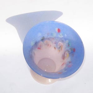 May include: A decorative glass vase with a light blue rim and a pale pink interior. The vase has colorful accents and a flared shape. The vase is sitting on a white surface.