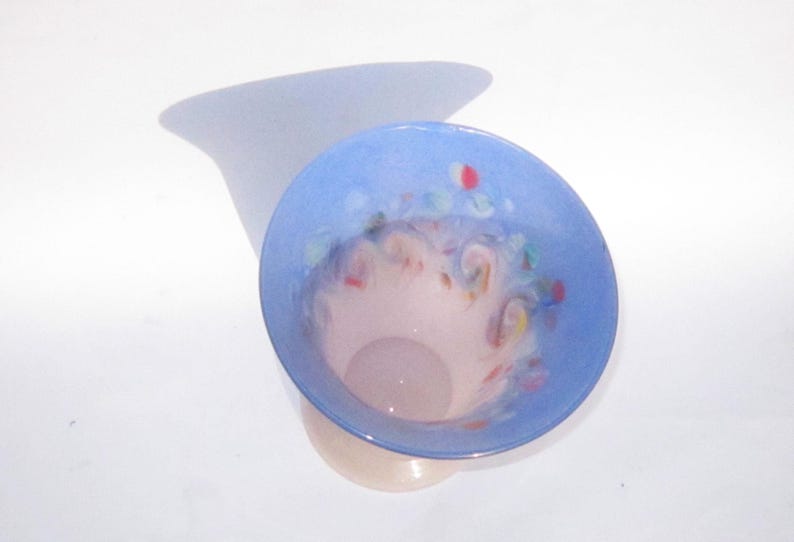 May include: A decorative glass vase with a light blue rim and a pale pink interior. The vase has colorful accents and a flared shape. The vase is sitting on a white surface.