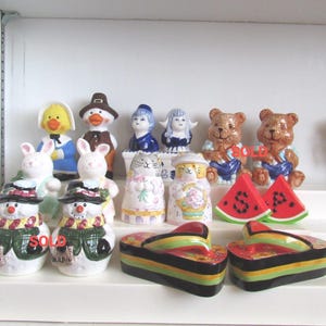 May include: A collection of ceramic salt and pepper shakers featuring various figures including snowmen, rabbits, bears, cats, people, and watermelon slices. Some items are marked with the word SOLD in red text on their surfaces.