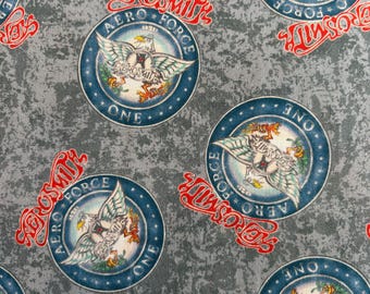 May include: A gray fabric featuring a repeating pattern of circular blue Aero Force One emblems with a central eagle graphic and the text Aerosmith in red script lettering. The background has a mottled, textured appearance in shades of gray.