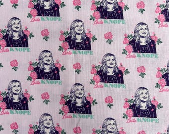 May include: A light pink fabric featuring a repeating pattern of a woman's portrait in a dark blazer, accompanied by pink roses with green leaves and the text "Leslie Knope" printed in a teal sans-serif font below each portrait.