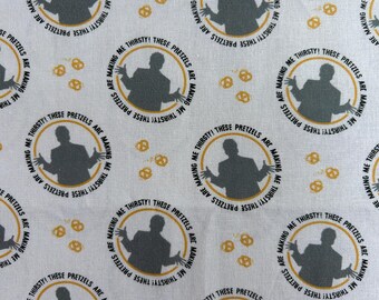 May include: A white fabric featuring a repeating pattern of dark gray silhouettes of a person gesturing within a gold circular border. The text "THESE PRETZELS ARE MAKING ME THIRSTY!" is printed in black along the circular border, interspersed with small gold pretzel icons.