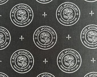 May include: A black fabric featuring a repeating white pattern of circular logos. Each logo contains a central rose illustration surrounded by the text Rose Apothecary and Handcrafted With Care. Small white fleur-de-lis symbols are placed between the logos.