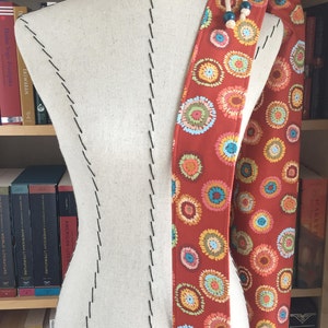 Puede incluir: Una bolsa de tela roja con cierre de cord&oacute;n y un patr&oacute;n circular colorido. La bolsa est&aacute; colgada de un maniqu&iacute; blanco.