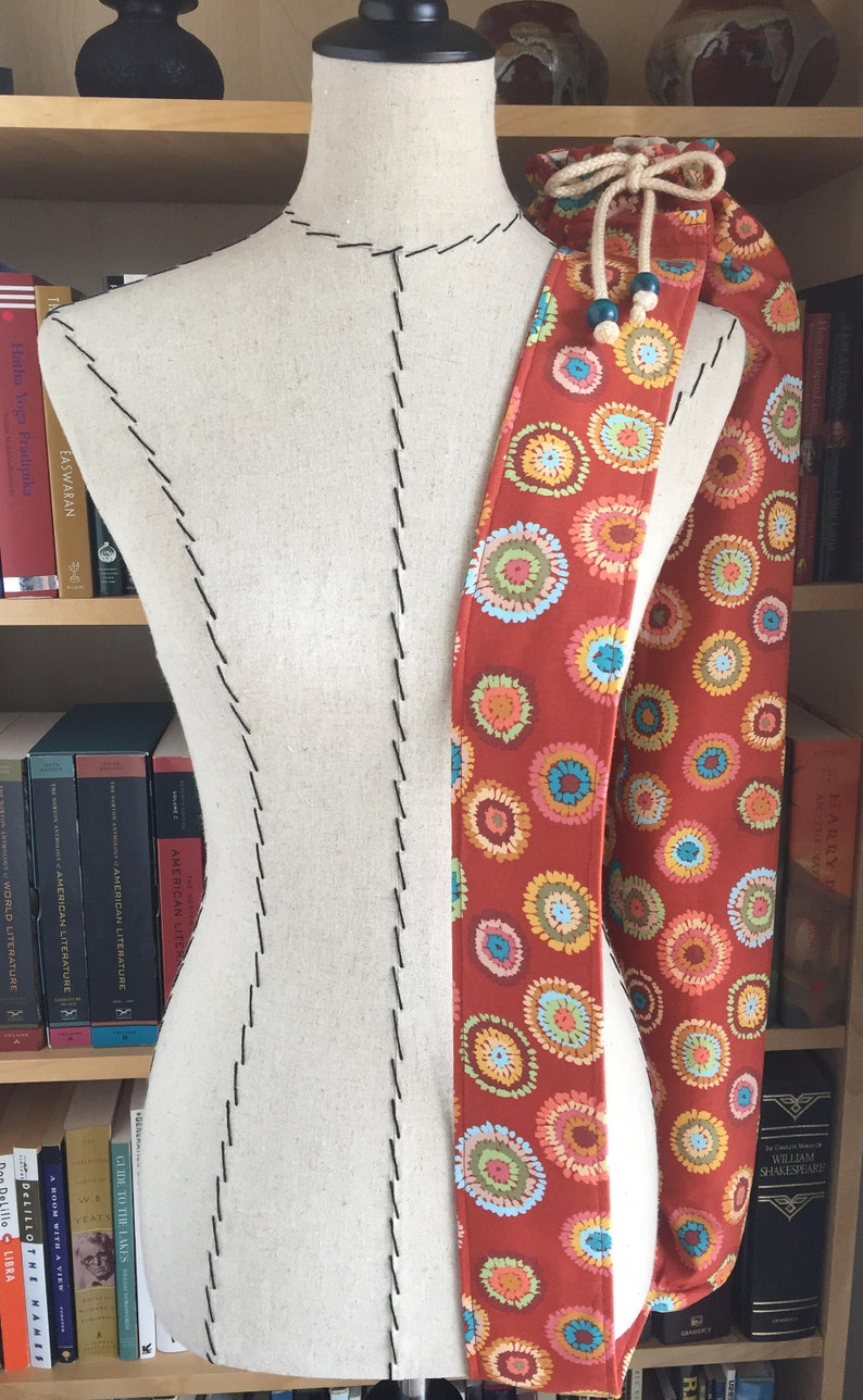 Puede incluir: Una bolsa de tela roja con cierre de cord&oacute;n y un patr&oacute;n circular colorido. La bolsa est&aacute; colgada de un maniqu&iacute; blanco.