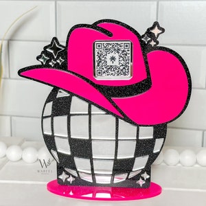May include: A pink glitter cowboy hat with a black and silver disco ball. The hat has a black band with a QR code on it. The disco ball is on a pink glitter base.