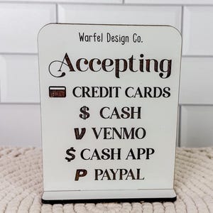 May include: A white wooden sign with the text "Warfel Design Co. Accepting" and payment methods: credit cards, cash, Venmo, Cash App, and PayPal. The sign is rectangular with a black base and is displayed on a beige surface.