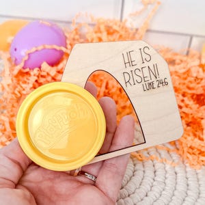 May include: A wooden arch with the text "HE IS RISEN!" and "LUKE 24:6" is held next to a yellow Play-Doh container. Easter eggs in shades of orange, purple, pink, and yellow are in the background, along with orange paper filler.