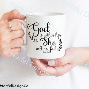 May include: White ceramic mug with the text "God is within her She will not fail" in black script, accented with leaf designs. The mug is held by a person. The mug has a handle and is a standard size.
