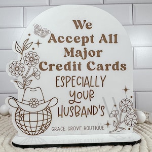 May include: White wooden sign with a brown wood-burned design. The sign reads "We Accept All Major Credit Cards Especially Your Husband's" with a butterfly and floral designs. The sign also has the text "Grace Grove Boutique" at the bottom.
