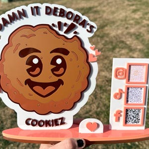 May include: A decorative cookie-shaped sign with the text "Damn It Debora's Cookiez". The sign features a smiling cookie face and social media QR codes. The sign is primarily brown, white, and orange.