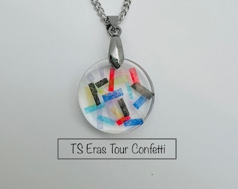 May include: A silver chain necklace with a round pendant featuring colorful confetti inside. The pendant has a clear background and the text "TS Eras Tour Confetti" is printed below the pendant.