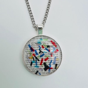May include: A silver chain necklace with a round pendant featuring a white background with the text "Taylor Swift" repeated many times. The text is covered with colorful confetti.