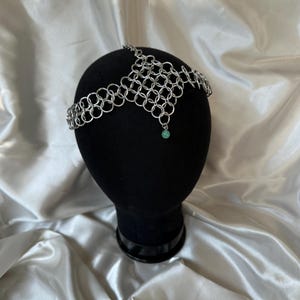 May include: A silver chainmail headpiece with a central design and a small, round, green bead hanging from the center. The headpiece is displayed on a black mannequin head against a white satin background.