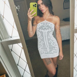 Op de afbeelding: Een witte halter-stijl gehaakte jurk met een geometrisch patroon. De jurk wordt gedragen met zwarte laarzen. De afbeelding is een spiegel selfie, die de textuur en pasvorm van de jurk laat zien. De jurk is knielang.