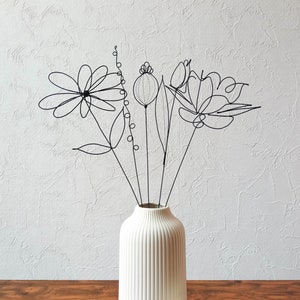 Peut inclure: Un vase en céramique blanche avec des nervures verticales contient un bouquet de fleurs en fil de fer noir. Les fleurs sont des représentations stylisées d'un coquelicot, d'une marguerite et d'une tulipe.