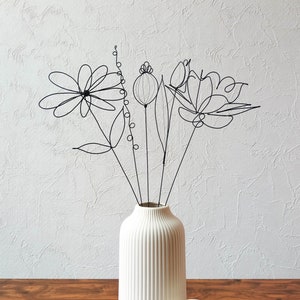 May include: A white, ribbed ceramic vase displays a collection of black wire flowers. The floral arrangement features a daisy, a poppy, and a tulip, each with a minimalist design. The vase rests on a wooden surface against a textured white wall.