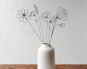 Puede incluir: Un jarrón de cerámica blanco con nervaduras verticales contiene un ramo de flores de alambre negro. Las flores son representaciones estilizadas de diferentes flores, incluyendo margaritas, amapolas y mariposas.