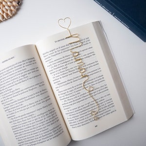 Può includere: Un segnalibro in filo dorato a forma di cuore con la parola "Hermione" scritta in corsivo. Il segnalibro è posizionato su un libro aperto con testo in francese.