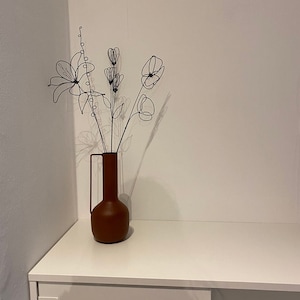 Puede incluir: Un jarr&oacute;n de terracota con una base ancha y un cuello estrecho contiene un ramo de flores de alambre. Las flores est&aacute;n hechas de alambre negro y tienen forma de lirios, amapolas y otras flores.