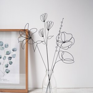 Puede incluir: Escultura de flores de alambre negro en un jarr&oacute;n de vidrio transparente. Las flores son estilizadas y abstractas, con l&iacute;neas y curvas delicadas. El jarr&oacute;n es simple y elegante, con una base redonda y un cuello estrecho.