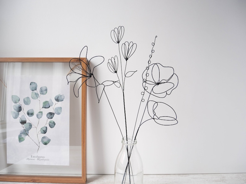 Puede incluir: Escultura de flores de alambre negro en un jarr&oacute;n de vidrio transparente. Las flores son estilizadas y abstractas, con l&iacute;neas y curvas delicadas. El jarr&oacute;n es simple y elegante, con una base redonda y un cuello estrecho.