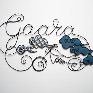 Op de afbeelding: Draadbeeld van de naam "Gaara" met gestileerde wolken en een motorfietsontwerp. De wolken zijn gemaakt van stof met een blauw-wit patroon. De motorfiets is gemaakt van zwart draad.