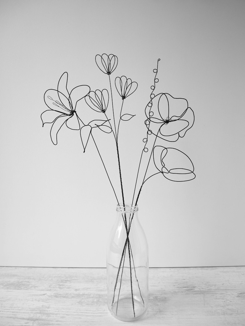 Puede incluir: Una escultura de flores de alambre negro en un jarr&oacute;n de vidrio transparente. Las flores son estilizadas e incluyen lirios, amapolas y otras flores.