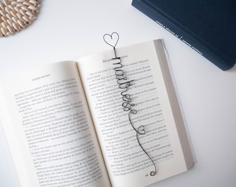 Puede incluir: Un marcapáginas de metal negro en forma de corazón con la palabra "MARVELOUS" escrita en cursiva. El marcapáginas está colocado en un libro abierto con texto en francés.