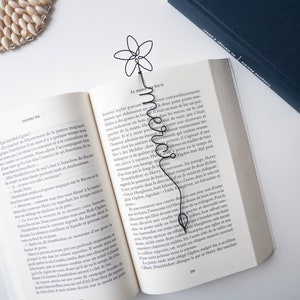 Op de afbeelding: Een zwarte draad bladwijzer in de vorm van een bloem met een stengel die zich krult tot het woord "Love". De bladwijzer is geplaatst in een open boek met witte pagina's en zwarte tekst. Het boek is getiteld "La Magie" en de tekst op de pagina is in het Frans.